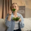 Evde koltukta otururken cam kaseden yeşil salata yiyen kadın
