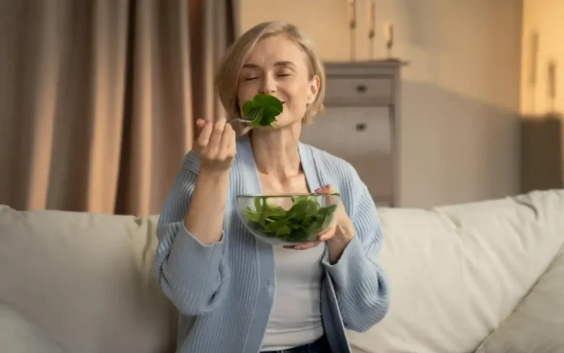 Evde koltukta otururken cam kaseden yeşil salata yiyen kadın